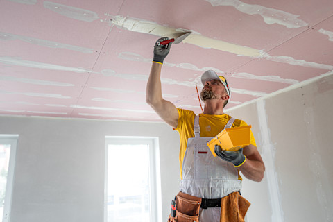 Een bouwvakker is bezig met het stucen van een plafond in een kamer. Hij draagt een gele T-shirt, een witte overall en een witte pet. In zijn rechterhand houdt hij een troffel waarmee hij het pleisterwerk op het plafond aanbrengt, terwijl hij in zijn linkerhand een gele bak met pleistermateriaal vasthoudt. Het plafond is bedekt met roze gipsplaten, waarvan de naden worden afgedekt met pleister. De kamer is nog in aanbouw, met onbewerkte muren en ramen die natuurlijk licht binnenlaten. De bouwvakker werkt geconcentreerd om het plafond glad af te werken.