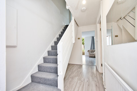 De afbeelding toont een hal in een huis met een trap die naar boven leidt. De trap is bekleed met grijze vloerbedekking en de muren zijn wit geschilderd. Op de vloer van de gang ligt een lichtgekleurde houten vloer. Aan de rechterkant van de gang is een grote spiegel aan de muur bevestigd, wat de ruimte groter laat lijken. In de verte is een lichte kamer zichtbaar, waarschijnlijk de woonkamer, met een bank en een raam waardoor daglicht naar binnen valt. De inrichting van de gang oogt strak en modern.