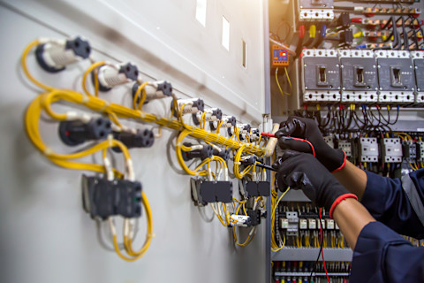 Een elektricien, gekleed in donkere werkkleding en zwarte handschoenen, werkt aan de bedrading in een elektrische kast. De kast is gevuld met gele stroomkabels en verschillende elektrische componenten. De elektricien gebruikt gereedschap om de bedrading aan te passen of te controleren. Op de achtergrond zijn meer schakelaars en bedrading te zien die deel uitmaken van een complex elektrisch systeem. De werkplek is goed verlicht, wat de details van het elektrische paneel duidelijk zichtbaar maakt. De scène toont een technisch en nauwkeurig werkproces in een industriële omgeving.