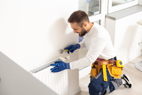 Op de afbeelding is een installateur te zien die bezig is met het afstellen of repareren van een radiator. Hij draagt blauwe werkhandschoenen en gebruikt een verstelbare moersleutel om aan de radiator te werken. De installateur heeft ook een gereedschapsriem met verschillende hulpmiddelen om zijn werk efficiënt uit te voeren. Het lijkt erop dat hij werkt in een moderne woning, met een lichte en schone omgeving. Dit beeld illustreert het onderhoud van verwarmingssystemen in huis.