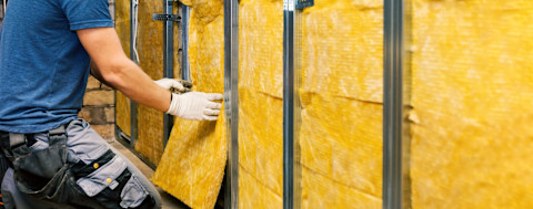 Twee handen die gele glaswol aanbrengen aan een muur