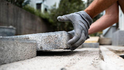 Stratenmaker met werkhandschoenen plaatst zorgvuldig een grijze betontegel tijdens het bestraten van een terras.
