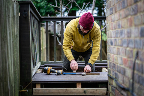 Een man in een gele jas en een rode muts is bezig met het plaatsen of vervangen van vlonders in een tuin. Hij knielt op een houten platform en gebruikt een hamer en andere gereedschappen die op de grond liggen, zoals een boormachine. De omgeving toont een houten hek aan de ene kant en een bakstenen muur aan de andere kant. Op de achtergrond zijn bomen en struiken zichtbaar.