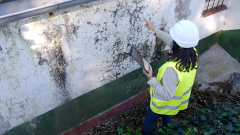 Een bouwkundig inspecteur bekijkt de donkere plekken op wand van een oudere woning. Ze maakt aantekeningen tijdens de inspectie.