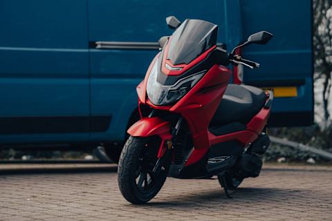 Rode maxi-scooter geparkeerd op straat, klaar voor aflevering aan nieuwe eigenaar.
