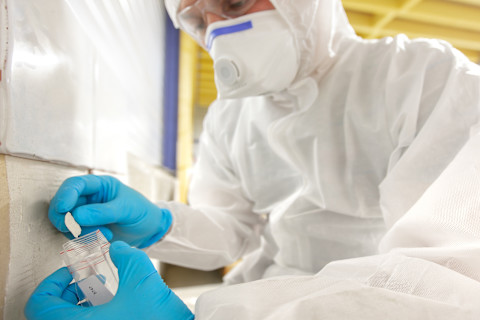 Op de foto zie je een persoon in een volledig witte beschermende pak, een masker en handschoenen. De persoon neemt voorzichtig een monster van een materiaal, waarschijnlijk voor analyse of inspectie. Het dragen van deze beschermende uitrusting suggereert dat de omgeving mogelijk schadelijke stoffen bevat, zoals asbest of chemische materialen. Het verzamelen van monsters zoals op de afbeelding wordt vaak gedaan in situaties waar veiligheid en precisie essentieel zijn.