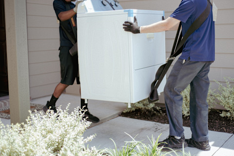 Twee verhuizers dragen een wasmachine naar de voordeur van een nieuw huis tijdens een verhuizing.