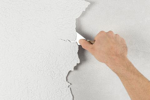 Hand verwijdert pleisterlaag van muur met een plamuurmes.