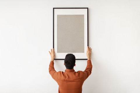  Een man hangt met beide handen een langwerpig leeg schilderij op aan een muur