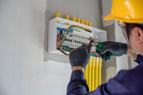 Een elektricien met een gele helm voert werkzaamheden uit bij een bedraad kastje
