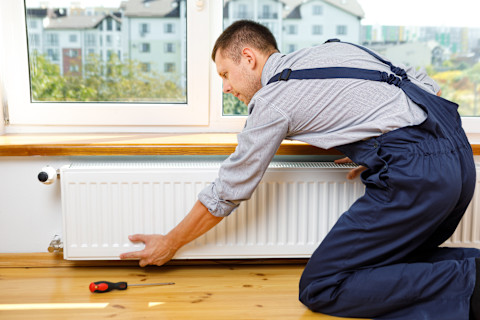 Een monteur, gekleed in een blauwe overall, knielt op de grond en is bezig met het verwijderen van een radiator in een woning. Er ligt gereedschap op de houten vloer.