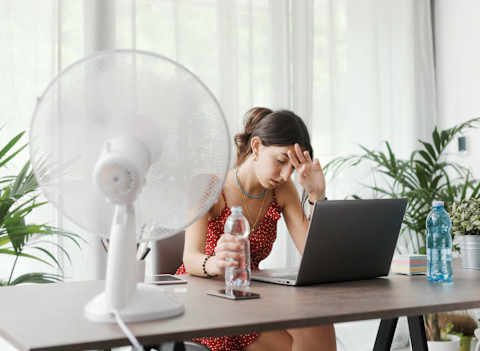 Vrouw zit uitgeput aan een bureau tijdens een warme zomerdag thuis, ventilator draait op volle kracht.