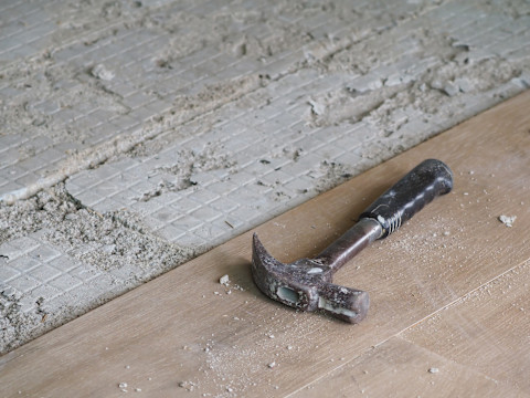 Een hamer ligt op een houten plank, met op de achtergrond een deels verwijderde vloer, met zicht op de ondervloer en wat puin.