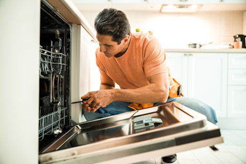 Technicus maakt met een schroevendraaier een reparatie aan een vaatwasser onder het aanrecht.