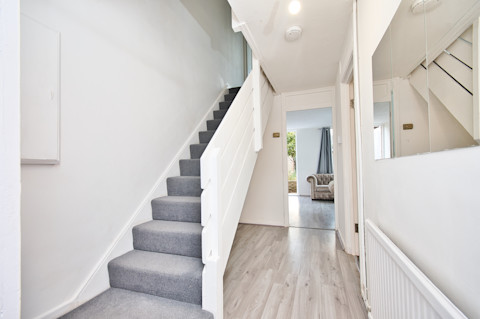 De afbeelding toont een hal in een huis met een trap die naar boven leidt. De trap is bekleed met grijze vloerbedekking en de muren zijn wit geschilderd. Op de vloer van de gang ligt een lichtgekleurde houten vloer. Aan de rechterkant van de gang is een grote spiegel aan de muur bevestigd, wat de ruimte groter laat lijken. In de verte is een lichte kamer zichtbaar, waarschijnlijk de woonkamer, met een bank en een raam waardoor daglicht naar binnen valt. De inrichting van de gang oogt strak en modern.
