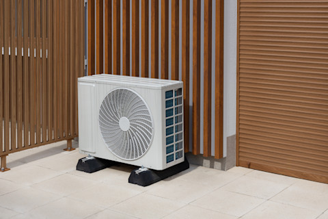 Op de foto is een airco-unit te zien die buiten een gebouw is geplaatst. De unit is wit en staat op zwarte steunen om hem van de grond te tillen. De achtergrond bestaat uit houten lamellen en een rolluik die beide een modern en strak ontwerp geven aan de ruimte. Deze opstelling zorgt ervoor dat de airco-unit goed geventileerd kan worden en tegelijkertijd mooi geïntegreerd is in de buitenomgeving.