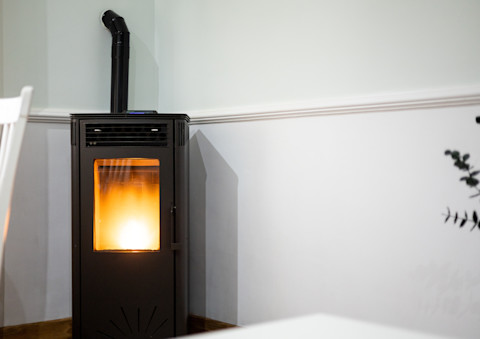 Een moderne pelletkachel staat in een hoek van een kamer, met een zachte, warme gloed die door het glazen raam aan de voorkant zichtbaar is. De kachel is zwart en heeft een eenvoudige, strakke vormgeving. De rookafvoer loopt via een zwarte pijp naar boven en verdwijnt in de muur. De muren van de kamer zijn licht van kleur, en er is een witte lambrisering zichtbaar. De omgeving ziet er netjes en rustig uit, en de kachel creëert een gezellige sfeer in de ruimte.