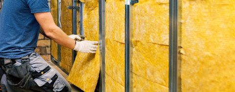 Twee handen die gele glaswol aanbrengen aan een muur