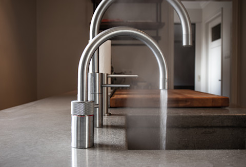 Een close-up van een moderne keuken met een Quooker-kraan die kokend water uitspuit. De kraan is van geborsteld staal en heeft een gebogen uitloop. Het kokende water komt in een gootsteen terecht, en er is lichte stoom zichtbaar. Naast de Quooker-kraan is een tweede standaard keukenkraan te zien. Het aanrecht is gemaakt van een glad, grijs materiaal, waarschijnlijk steen of beton. Op de achtergrond is een houten snijplank op het aanrecht en een deel van de keuken met planken en een deur te zien.