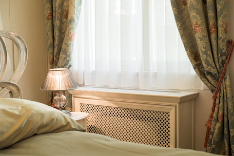 Houten radiatorombouw in lichte woonkamer – een houten ombouw met roosterpatroon rond een radiator, geplaatst tegen een witte muur.