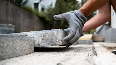 Stratenmaker met werkhandschoenen plaatst zorgvuldig een grijze betontegel tijdens het bestraten van een terras.