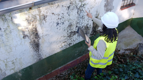 Een bouwkundig inspecteur bekijkt de donkere plekken op wand van een oudere woning. Ze maakt aantekeningen tijdens de inspectie.