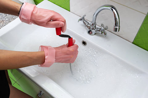 Vrouw met rubberen handschoenen probeert een verstopte afvoer in de keuken te ontstoppen.