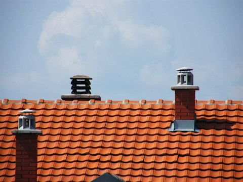 Een dak met nieuwe oranje dakpannen en drie schoorstenen, twee bakstenen en één metalen. De dakpannen zijn recent vervangen en hebben een glanzende afwerking. De lucht op de achtergrond is blauw met enkele witte wolken, wat een helder en zonnig weer suggereert. Dak vervanging, dakrenovatie