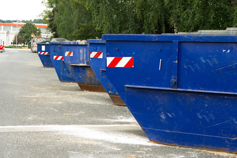 Op de afbeelding staan meerdere blauwe afvalcontainers op een rij, klaar voor gebruik. Ze zijn voorzien van reflecterende rood-witte waarschuwingsstickers, waarschijnlijk om ze zichtbaar te maken voor verkeer. De containers bevinden zich op een parkeerplaats of terrein, omringd door bomen en gebouwen in de verte. Het lijkt erop dat deze containers worden gebruikt voor grofvuil of bouwafval.