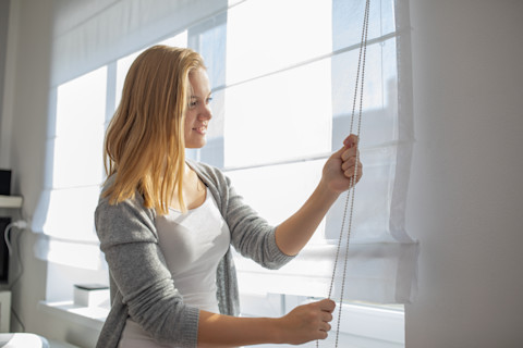 Vrouw hangt rolgordijn op bij raam in modern interieur, met natuurlijke lichtinval.