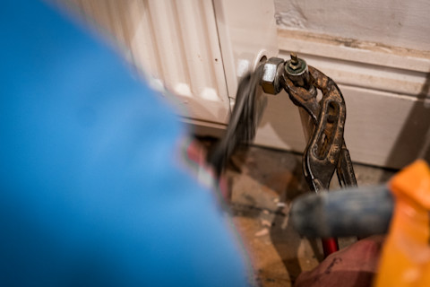 Een close-up van een monteur die bezig is met het aansluiten van een radiator. De monteur gebruikt een moersleutel om de koppeling tussen de radiator en de pijp aan te draaien. De omgeving lijkt een beetje stoffig en de vloer heeft een werkgebied met gereedschap.