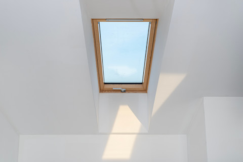 Een dakraam in een zolderkamer in ruwbouwstaat met zichtbare dakspanten en daglicht dat door het raam naar binnen valt.