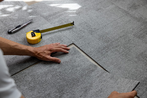 Op de afbeelding zie je iemand bezig met het leggen van tapijttegels. De persoon gebruikt een meetlint en een stanleymes om de tegels nauwkeurig op maat te snijden en passend te maken. De grijskleurige tegels worden zorgvuldig uitgelijnd om een nette afwerking te garanderen. Dit soort werk vereist precisie, vooral bij het snijden en plaatsen van de tegels, zodat ze strak tegen elkaar aan liggen en een naadloze vloer vormen.
