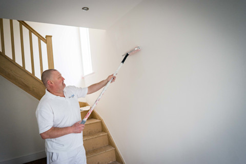Op de afbeelding is een schilder bezig met het schilderen van een muur. Hij gebruikt een verfroller met een lange steel om de verf gelijkmatig aan te brengen op de witte muur. De schilder staat in een huis bij een trap, en draagt een witte werkoutfit, wat typisch is voor een professionele schilder. De houten trap achter hem leidt naar boven, en de ruimte oogt licht door het natuurlijke licht dat binnenvalt.