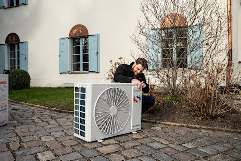 Een technicus is bezig met de installatie van een airco-unit buiten een woning. De man knielt naast de airco-unit en gebruikt gereedschap om aanpassingen te maken. De unit staat op een betegeld oppervlak naast een tuin met kale struiken. Op de achtergrond is een traditionele woning te zien met crèmekleurige muren en blauwe luiken voor de ramen. De ramen hebben een halfronde decoratie aan de bovenkant. De scene vindt plaats op een bewolkte dag, wat te zien is aan het gedempte licht.