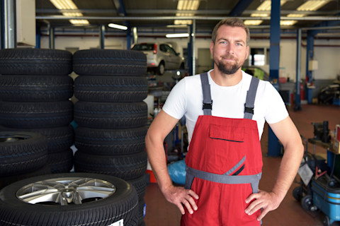 Glimlachende automonteur staat in de werkplaats klaar voor onderhoud of reparatie aan een auto.