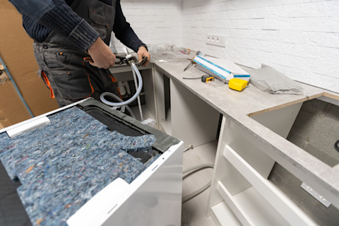 Een installateur is bezig met het aansluiten van een vaatwasser in een keuken. De persoon, gekleed in donkere werkkleding, gebruikt een tang om een waterslang aan te sluiten op de waterleidingen onder het aanrecht. Op het aanrecht liggen verschillende gereedschappen en materialen, waaronder een rol afplaktape, een meetlint en een gele waterpas. De keuken is nog in aanbouw, zoals te zien is aan de onafgewerkte kasten en het ontbrekende aanrechtblad aan de rechterkant. De omgeving suggereert een werkplaats of een renovatieproject in een woning.
