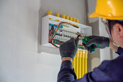 Een elektricien met een gele helm voert werkzaamheden uit bij een bedraad kastje
