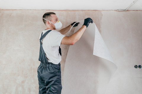 Behanger in overal met handschoenen en beschermend gezichtsmasker is bezig met het aanbrengen van renovlies behang op de muren