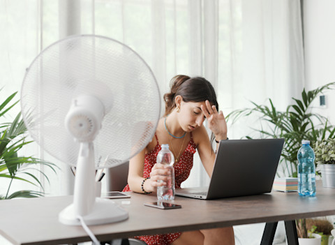 Vrouw zit uitgeput aan een bureau tijdens een warme zomerdag thuis, ventilator draait op volle kracht.