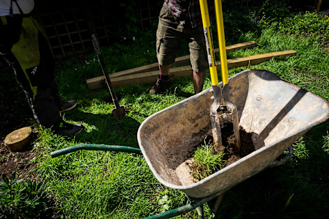 Twee personen zijn bezig met tuinwerkzaamheden. In het midden van de afbeelding staat een kruiwagen met aarde en gras, waarin een grondboor staat. Een van de personen draagt een groene tuinbroek en zwarte schoenen, terwijl de ander een korte broek en bruine werkschoenen draagt. Op de achtergrond liggen enkele houten balken op het gras. Het gras is groen en de tuin lijkt goed onderhouden.