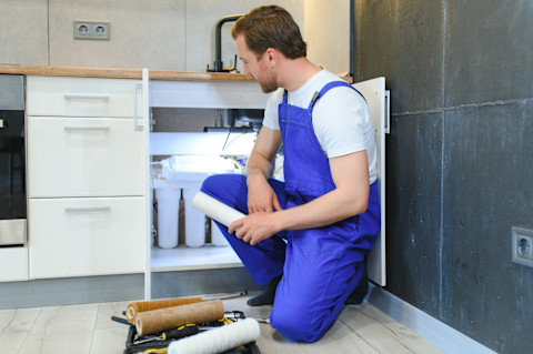Loodgieter vervangt patroonfilter van een waterfilterinstallatie onder de gootsteen, zichtbaar onderhoudswerk aan leidingsysteem.