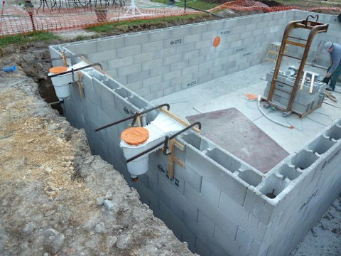 Een bouwplaats waar de fundering van een gebouw wordt aangelegd. De fundering bestaat uit betonnen blokken die zorgvuldig zijn geplaatst om een stevige basis te vormen. Aan de zijkant van de fundering zijn enkele bouwmaterialen en gereedschappen zichtbaar, waaronder emmers en stalen stangen. Een werker is bezig met de werkzaamheden binnen de fundering. De omgeving is afgebakend met oranje veiligheidsnetten.
