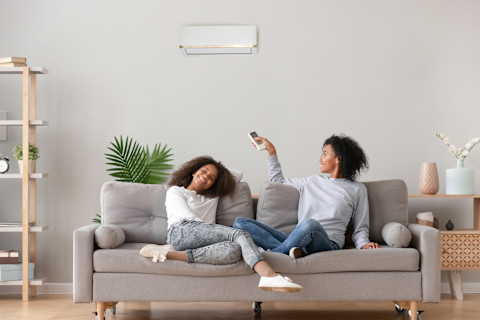 Meisje gewikkeld in een deken zet een airco aan of uit met een afstandsbediening, zittend op een bank in een warme woonkamer.