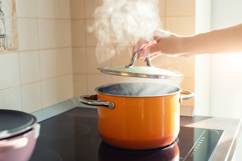 Vrouwelijke hand opent de deksel van een emaille pan op een inductiekookplaat in een moderne keuken.