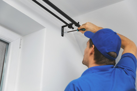 Klusjesman in blauwe kleding hangt een zwarte gordijnrails op aan een witte muur