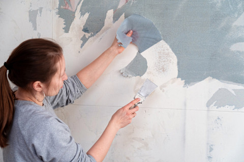 Vrouw verwijdert behang van een muur, zichtbaar halverwege het losmaken van de stroken.