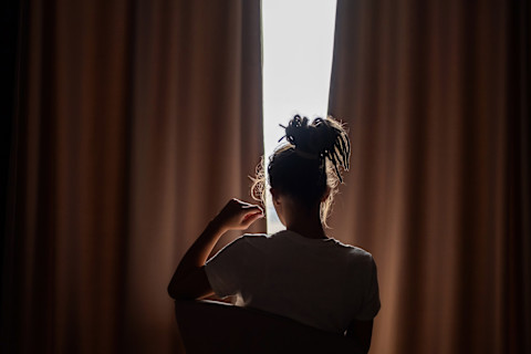 Persoon zit in silhouet voor een raam met gesloten rolgordijn dat licht filtert.