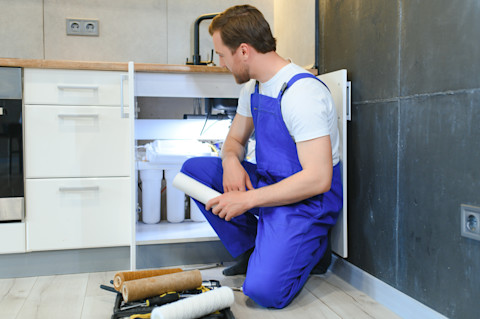 Loodgieter vervangt patroonfilter van een waterfilterinstallatie onder de gootsteen, zichtbaar onderhoudswerk aan leidingsysteem.