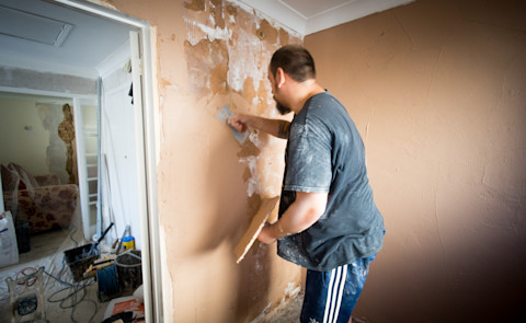 Een man is bezig met het gladstrijken van een muur in een woning. Hij gebruikt een plamuurmes en een stuk gipsplaat om de muur glad te maken en eventuele beschadigingen te repareren. De kamer is duidelijk in renovatie, met meubels bedekt met plastic en gereedschap en bouwmaterialen verspreid over de vloer. De muur waar de man aan werkt, toont plekken van oude verf en pleister die wordt bijgewerkt.